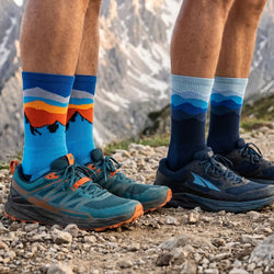 Two pairs of feet wearing colorful socks and running shoes on a rocky ground with mountains in the background.