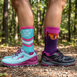 Person wearing colorful socks with 'Run Like a Girl' and nature-themed designs, standing on a forest path.