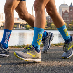 Two pairs of legs wearing blue running socks with 'Marathon' and 'Boston' text, running on a path by a body of water.