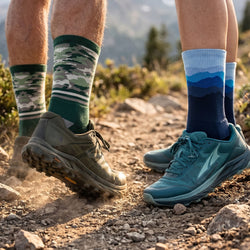 Two pairs of feet wearing colorful socks and running shoes on a rocky trail with a mountainous background.