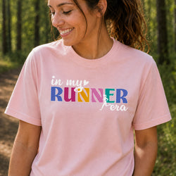 A woman wearing a light pink graphic t-shirt with the text 'in my RUNNER era' printed on the front, set against a blurred forest background.