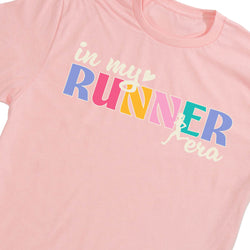 A close-up of a light pink crewneck t-shirt featuring a colorful graphic that reads 'in my RUNNER era' in pastel blue, pink, yellow, and teal block letters with cream script text.