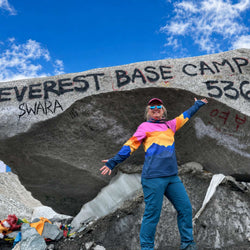 Person posing in front of a rock with 'Everest Base Camp 536' inscription against a blue sky.