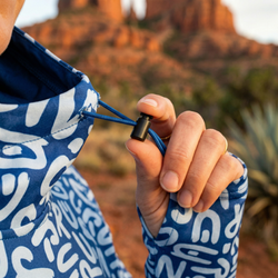 Person wearing a blue patterned jacket showing an adjustable toggle for a hood with a blurred desert landscape in the background