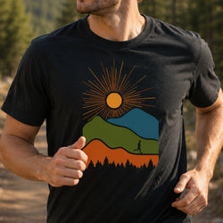 A man wearing a black t-shirt with a colorful mountain and sun graphic, photographed outdoors in a forest setting.
