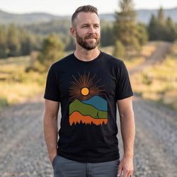 A man wearing a black t-shirt with a colorful mountain and sun graphic, standing on a gravel path in an outdoor mountain landscape.