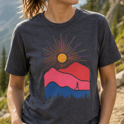 A woman wearing a dark heather gray t-shirt with a colorful graphic of a sun, pink and blue mountains, and a silhouette of a runner, set against a blurred outdoor mountain background.