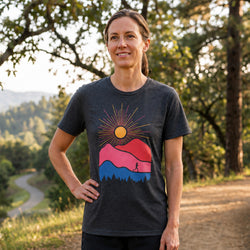 A woman standing outdoors on a dirt path wearing a dark heather gray graphic t-shirt with a colorful mountain and sun design, set against a blurred background of trees and a winding road.