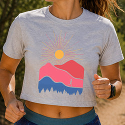 A woman wearing a light heather gray cropped t-shirt with a colorful mountain and sun graphic on the front, set against a blurred outdoor background.