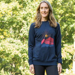 Woman wearing a navy blue sweatshirt with a red and pink mountain design, standing outdoors with greenery in the background.