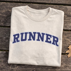 A folded off-white t-shirt with the word 'RUNNER' printed in navy blue varsity-style letters with a white outline, laid flat on a rustic dark wood plank surface.