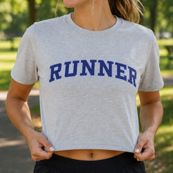 A woman wearing a light heather gray cropped t-shirt with the word 'RUNNER' printed in navy blue arched letters across the chest, set against a blurred green park background.