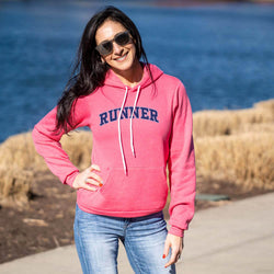 Woman wearing a pink hoodie with 'RUNNER' printed on it, standing by a body of water.