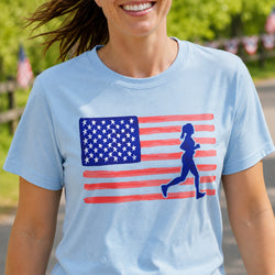 A woman wearing a light blue crewneck t-shirt with a graphic of the American flag and a blue silhouette of a running woman, set against a blurred outdoor background.
