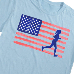 A close-up of a light blue t-shirt featuring a graphic print of a stylized American flag with a blue silhouette of a running woman.