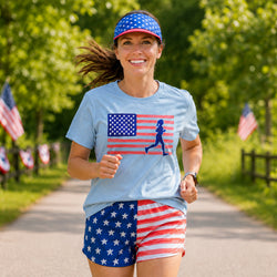 A woman running outdoors on a tree-lined path wearing a light blue t-shirt with a red, white, and blue American flag graphic, matching patriotic shorts, and a matching visor.