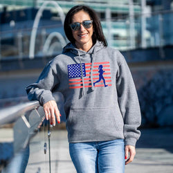 Woman wearing a gray hoodie with an American flag and running figure design, standing outdoors.