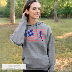 Woman wearing a gray hoodie with an American flag and running figure design, standing outdoors.
