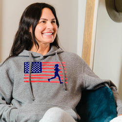 Woman wearing a gray hoodie with an American flag and running person design, sitting indoors.