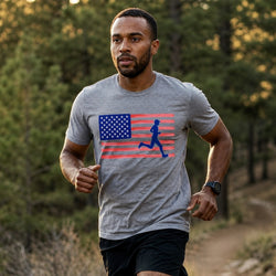 A man running outdoors on a wooded trail wearing a heather gray t-shirt with a graphic of an American flag and a blue running silhouette, paired with black shorts.