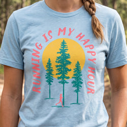 A person wearing a light blue graphic t-shirt with a forest and running theme. The graphic features a yellow sun, three green pine trees, a small pink runner silhouette, and the text 'RUNNING IS MY HAPPY HOUR' in pink.