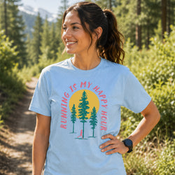 A woman wearing a light blue graphic t-shirt with a 'RUNNING IS MY HAPPY HOUR' design featuring pine trees and a yellow sun, standing on a forest path.