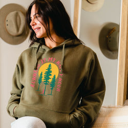 Woman wearing a green hoodie with nature-themed happy hour design, sitting in a room with hats on the wall.
