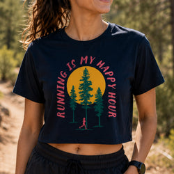 A woman wearing a navy blue cropped t-shirt with a graphic print that reads 'RUNNING IS MY HAPPY HOUR' in pink around a yellow sun and green trees. The background is a blurred outdoor forest path.