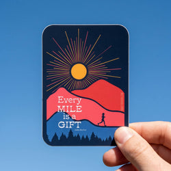 Sticker with sun and mountains design held by a hand against a blue sky