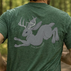 A man wearing a heather green t-shirt, viewed from behind, featuring a large gray graphic of a running rabbit with antlers and the text 'RUN DIFFERENT' printed on the back, set against a blurred forest background.