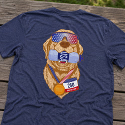 A navy blue heather t-shirt laid flat on a weathered wooden surface, featuring a graphic of a golden retriever wearing sunglasses with the American flag, a water bottle with 'USA' on it, a medal, and a race bib.