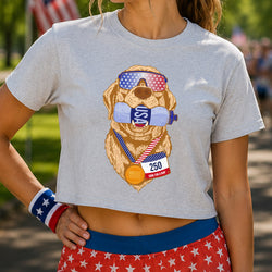 A woman wearing a light heather gray cropped t-shirt with a graphic of a golden retriever dog wearing sunglasses, a race bib, and a medal. The background is a blurred outdoor park setting.