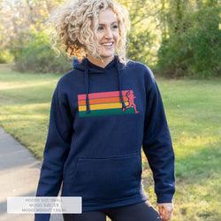 A woman outdoors wearing a navy blue hoodie with a colorful chest graphic showing a red, orange, yellow, and green striped background with a pink running figure. 