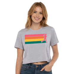 A woman wearing a heather gray cropped t-shirt with a pink silhouette of a runner over horizontal pink, orange, yellow, and green stripes, paired with blue jeans, against a plain white background.