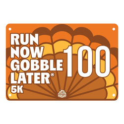 An orange and brown 5K race bib with the text 'RUN NOW GOBBLE LATER 100 5K' in white, featuring a stylized parachute graphic and a small 'Run Now Gobble Later' logo at the bottom center.