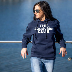 A woman wearing a navy blue hoodie with white text that reads 'THE Daily Run CLUB' and light blue jeans, standing outdoors against a blue water background.