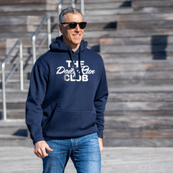 A man wearing a navy blue hoodie with 'THE Daily Run CLUB' printed in white across the chest, paired with blue jeans and black sunglasses, standing outdoors in front of wooden stadium-style steps.
