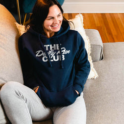 A woman sitting on a gray couch wearing a navy blue hoodie with white text that reads 'THE Daily Run CLUB' and light gray sweatpants in a living room setting.