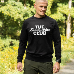 A man wearing a black long-sleeve crewneck sweatshirt with 'THE Daily Run CLUB' printed in white across the chest, standing outdoors on a path with green trees and yellow flowers in the background.
