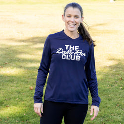 A woman standing outdoors on a grassy lawn wearing a navy blue long-sleeve athletic shirt with 'THE Daily Run CLUB' printed in white across the chest, paired with black leggings.