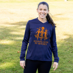 A woman standing outdoors on a grassy lawn wearing a navy long-sleeve V-neck shirt with an orange graphic of three witches and the text 'RUNNING WITH MY WITCHES' on the front.