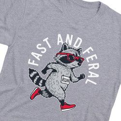 A close-up of a heather gray t-shirt featuring a cartoon raccoon running with red sunglasses, a white bib that says 'RUN', and the words 'FAST AND FERAL' printed in white around it.