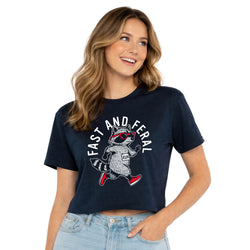 A woman wearing a navy blue cropped t-shirt with a graphic of a raccoon wearing red sunglasses and red sneakers, with the text 'FAST AND FERAL' arched above it. She is paired with light-wash blue jeans against a clean white background.