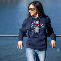 A woman wearing a navy blue hoodie with a cartoon raccoon graphic and the text 'FAST AND FERAL' stands outdoors near a railing with water in the background.