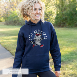A woman wearing a navy blue hoodie with a raccoon graphic and the words 'FAST AND FERAL' on the front, standing outdoors on a grassy path with trees in the background.