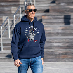 A man wearing a navy blue hoodie with a 'Fast and Feral' graphic on the chest, paired with blue jeans and sunglasses, standing outdoors in front of wooden stadium steps.