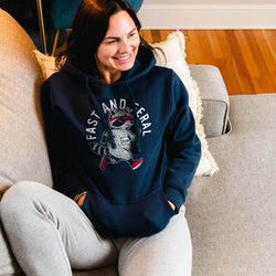 A woman sitting on a gray couch wearing a navy blue hoodie with a raccoon graphic and the words 'FAST AND FERAL' in white, paired with light gray sweatpants in a living room setting.