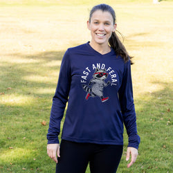 A woman standing outdoors on a grassy lawn wearing a navy blue long-sleeve athletic shirt with a raccoon graphic and the words 'FAST AND FERAL' printed on the front, paired with black leggings.