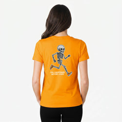 A woman viewed from behind wearing a bright orange short-sleeve T-shirt with a back graphic of a running skeleton and the text 'I'M A NIGHTMARE BEFORE A RUN'.