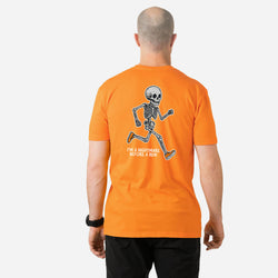A man viewed from behind wearing an orange t-shirt with a black and white running skeleton graphic and the text 'I'M A NIGHTMARE BEFORE A RUN' printed on the back, paired with black pants against a light gray background.